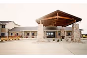 Exterior front entrance of a retirement community building featuring a large wooden porte-cochère supported by stone pillars and a paved driveway.