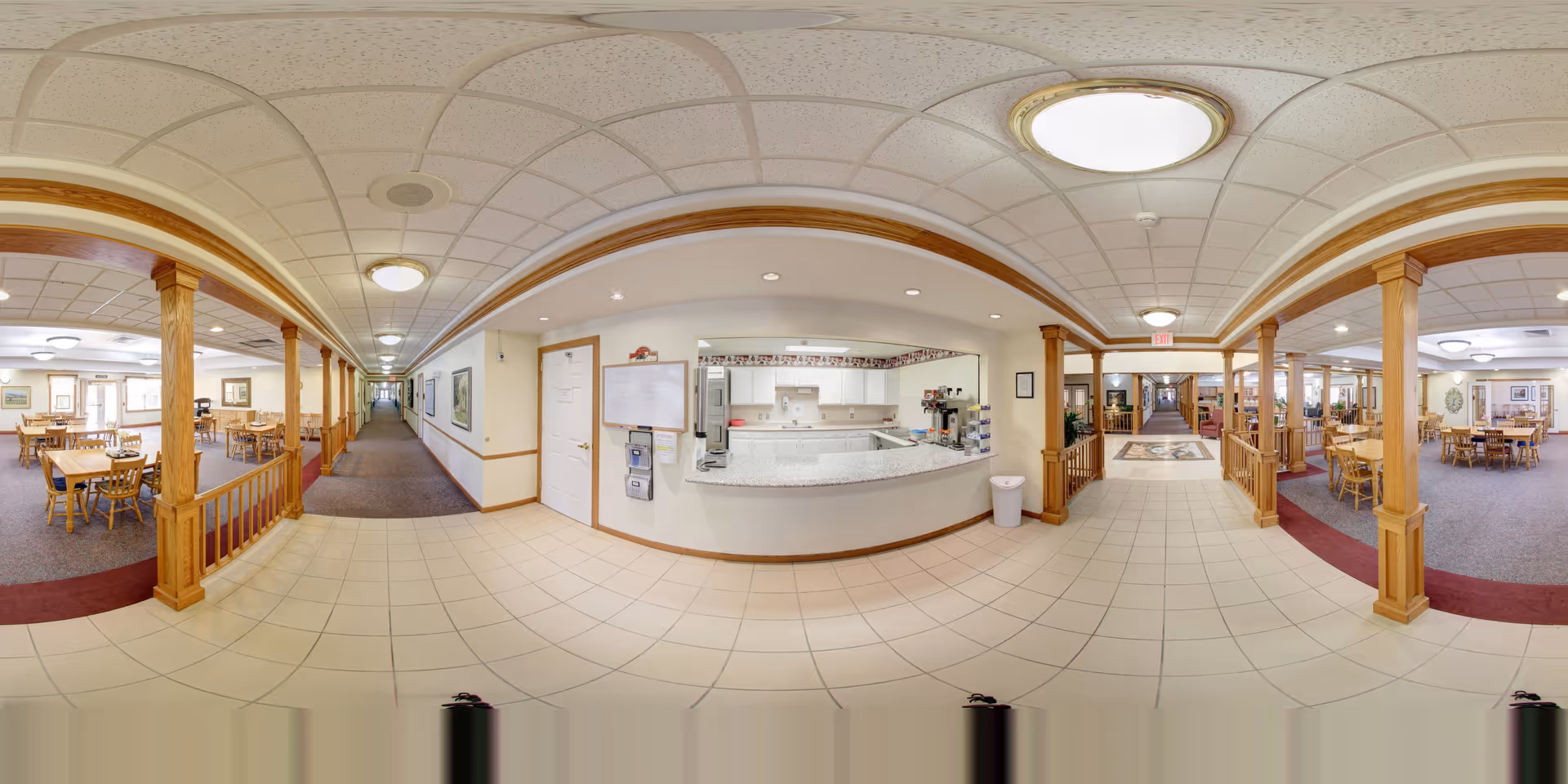Wide panoramic view of a senior living facility interior showing a central hallway with tiled floor and ceiling lights. On both sides of the hallway are dining areas with wooden tables and chairs, separated by wooden columns and railings. In the center is a kitchen service counter with white cabinets, a coffee machine, and a water dispenser. The space is well-lit with natural and artificial light.