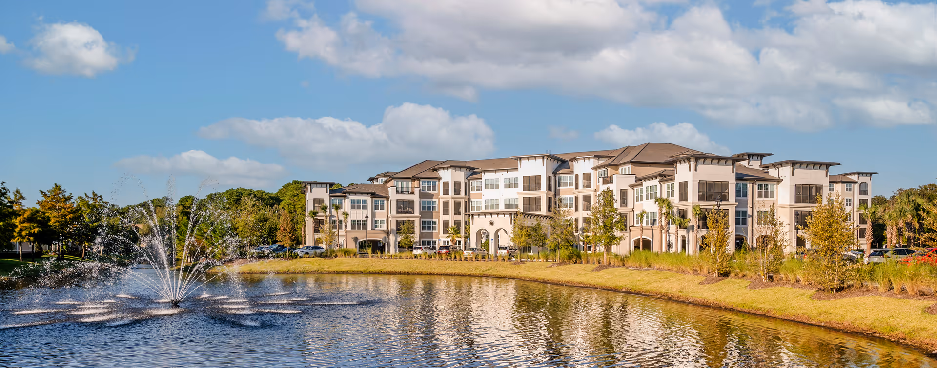 A large, multi-story senior living facility building named Fleet Landing is situated behind a pond with a water fountain. The building is surrounded by trees and landscaping under a partly cloudy blue sky.