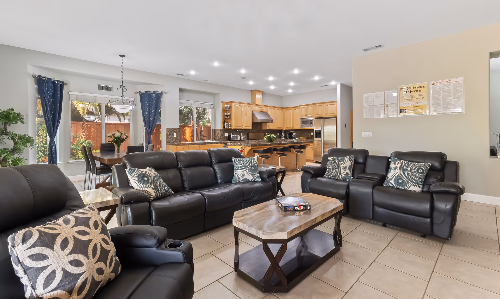 A spacious living room area with black leather sofas and patterned pillows, a wooden coffee table with a board game on it, and a dining area with a table and chairs near large windows with blue curtains. The background shows a kitchen with wooden cabinets, stainless steel appliances, and a breakfast bar with stools.