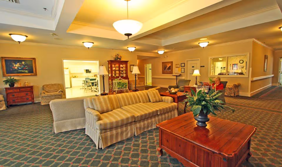 A spacious senior living facility common area with beige walls and patterned carpet. The room features a striped sofa, armchairs, wooden coffee tables with potted plants, and lamps. In the background, a woman is seated near a reception window. The area is well-lit with ceiling lights and has a cozy, welcoming atmosphere.