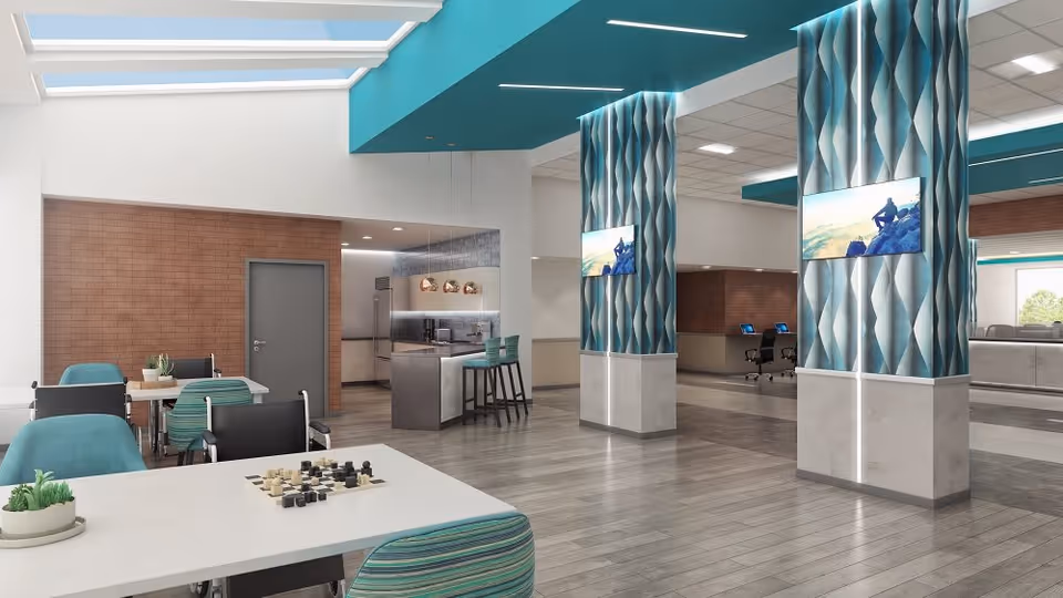 Bright, modern communal dining and activity area with tables and chairs, patterned columns with mounted TVs, and a small kitchen/bar.