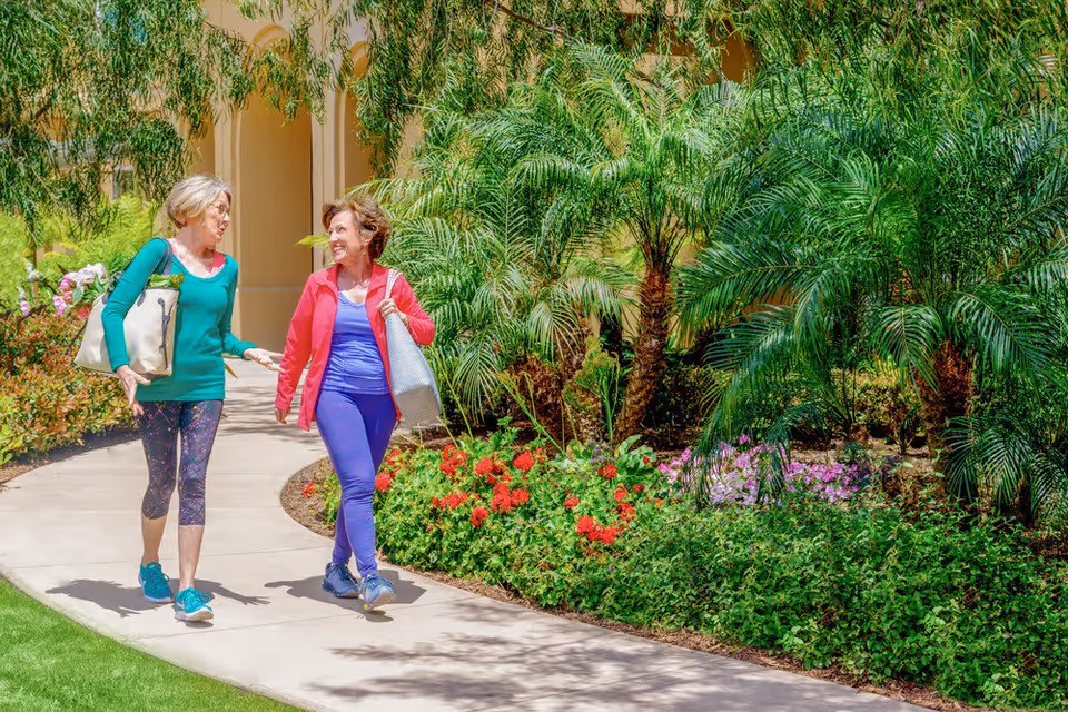 Two older women walking and talking on a curved garden pathway surrounded by lush green plants and colorful flowers at a senior living community.