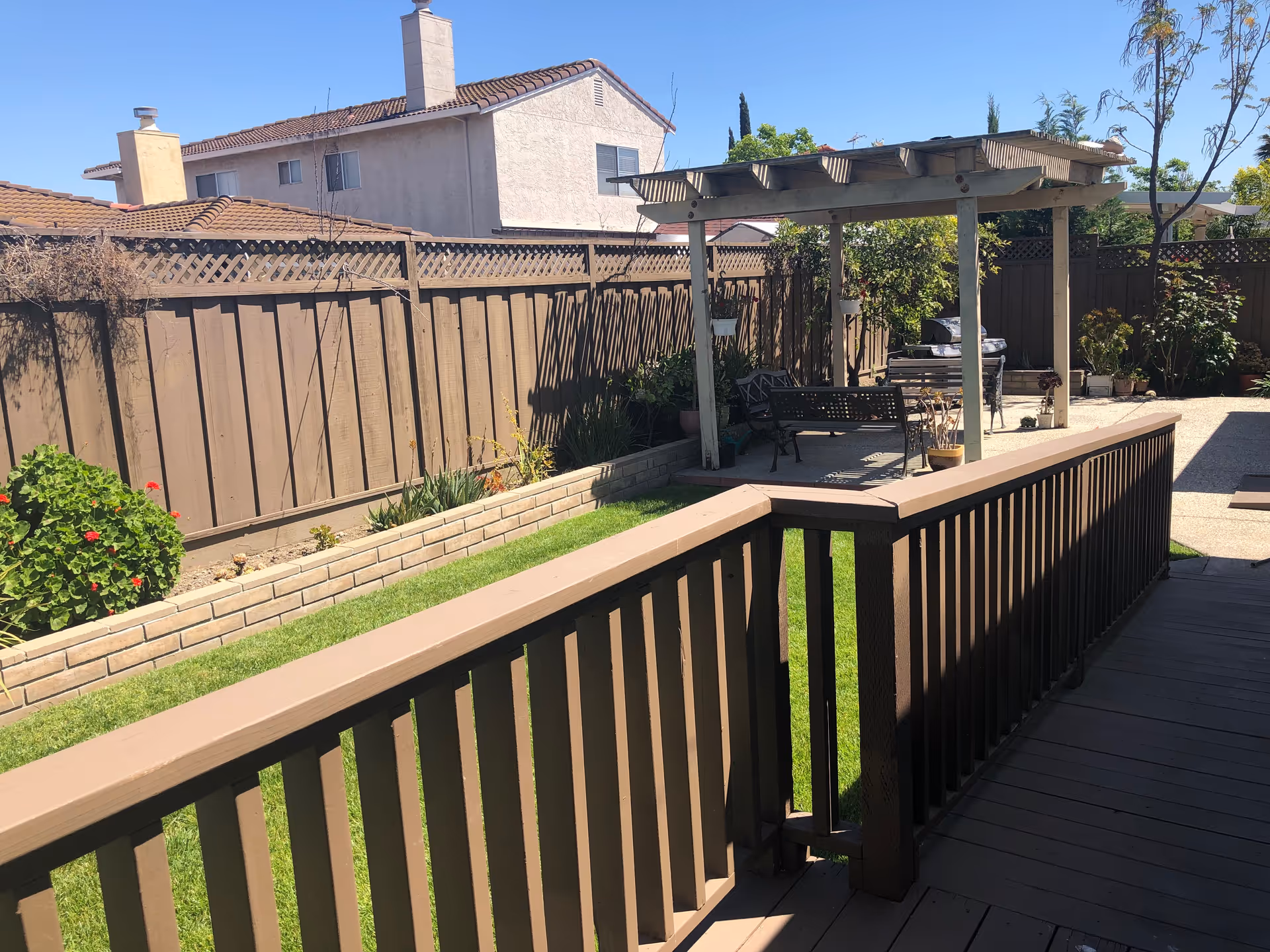 Outdoor area of a residential care facility featuring a wooden deck with railing, a small grassy lawn, a raised flower bed along a wooden fence, and a pergola with seating and a barbecue grill under it. The sky is clear and blue.