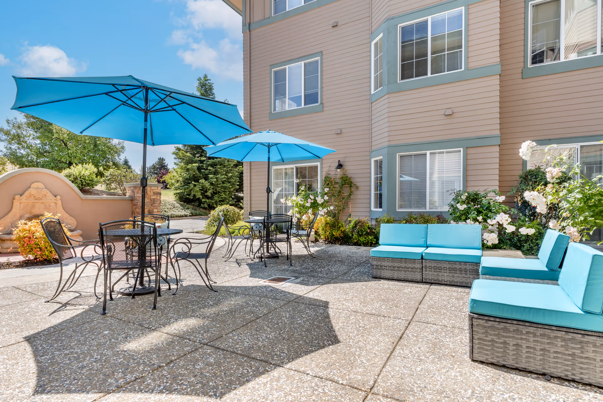 Outdoor patio area at Brookdale San Ramon featuring two black metal tables with matching chairs under bright blue umbrellas, and blue cushioned wicker seating along the building wall. The patio is surrounded by greenery and flowers, with a beige building and windows in the background under a partly cloudy sky.