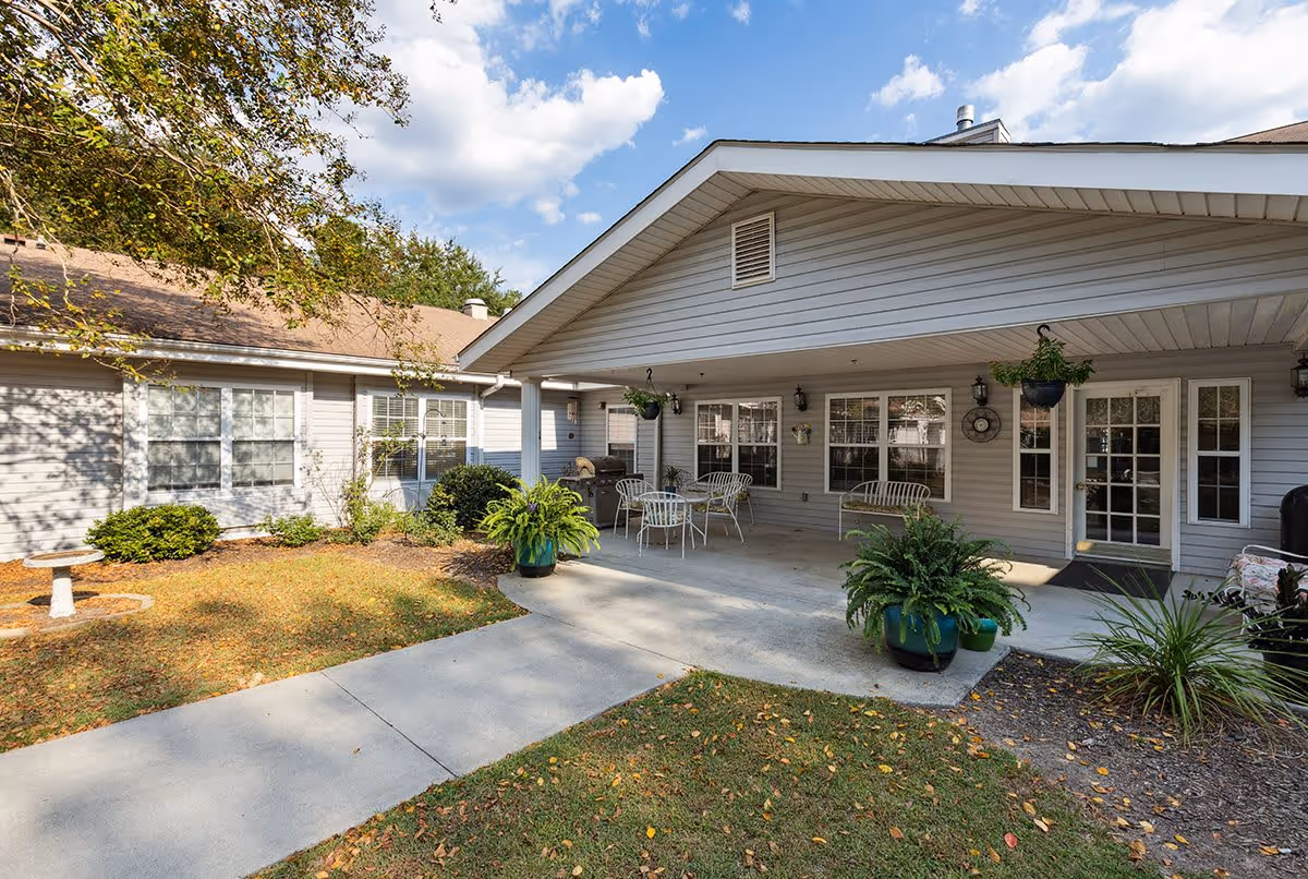 Outdoor patio area at Hitchcock Square with a covered seating space featuring metal chairs and tables, surrounded by potted plants and greenery. The building exterior is light gray with multiple windows and a door leading inside. The sky is partly cloudy.