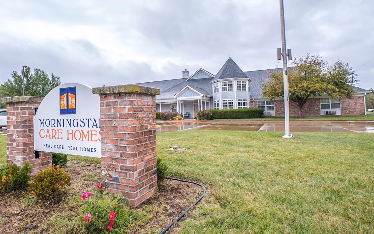 Exterior view of Morningstar Care Homes of Neodesha, showing a large brick and siding building with a turret-like feature and multiple windows. In the foreground, there is a brick sign with the facility's name and slogan, surrounded by grass and some small plants. The sky is overcast and the ground appears wet from recent rain.