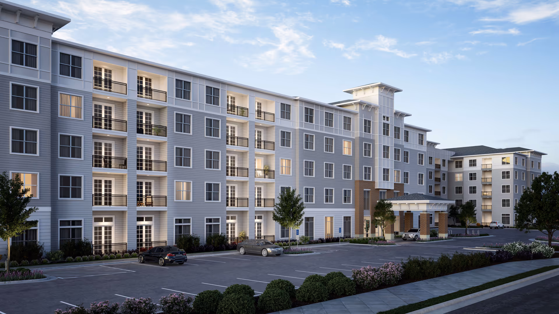 Exterior view of a modern multi-story senior living facility named Overture Chapel Hill, featuring balconies, large windows, a covered entrance, landscaped greenery, and a parking lot with several cars under a partly cloudy sky.