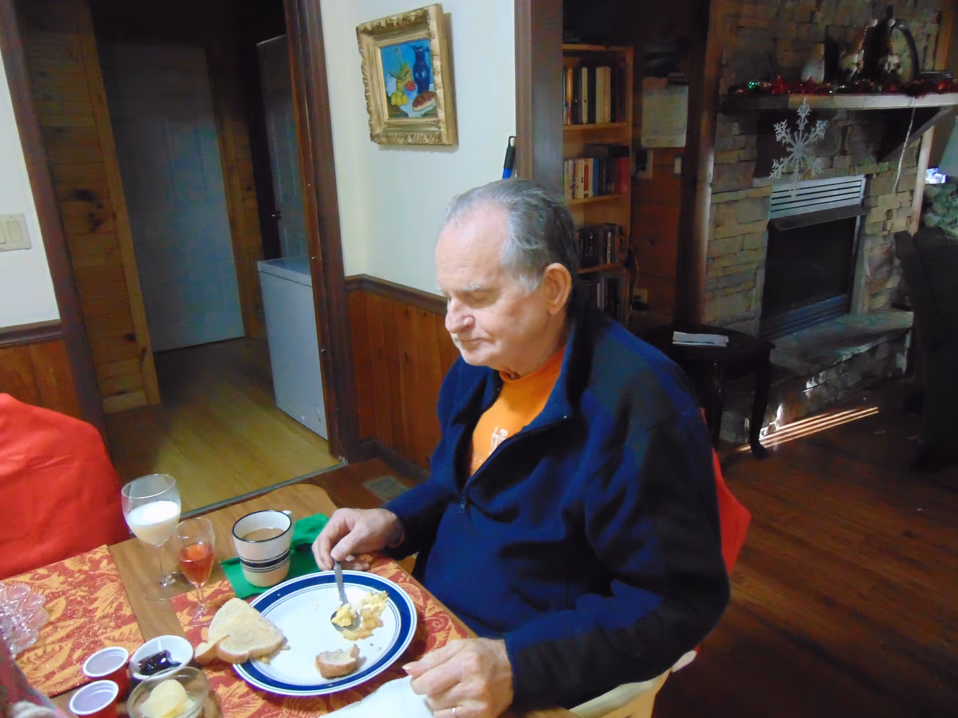 An elderly man sits at a dining table eating breakfast with plates, cups, and a glass of milk in a cozy room with a stone fireplace.