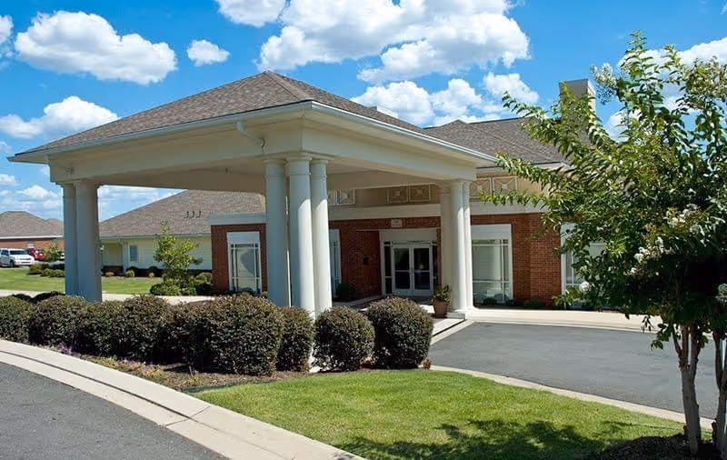 Entrance of Mt. Carmel Community Benton facility showing a covered drop-off area with white columns, brick exterior walls, and a driveway surrounded by green bushes and a tree under a blue sky with scattered clouds.