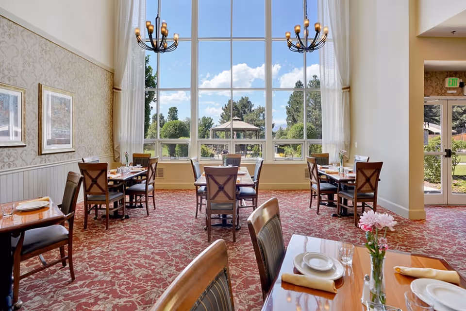 Bright dining room with multiple set tables and chairs, tall windows overlooking a garden gazebo, and chandeliers.