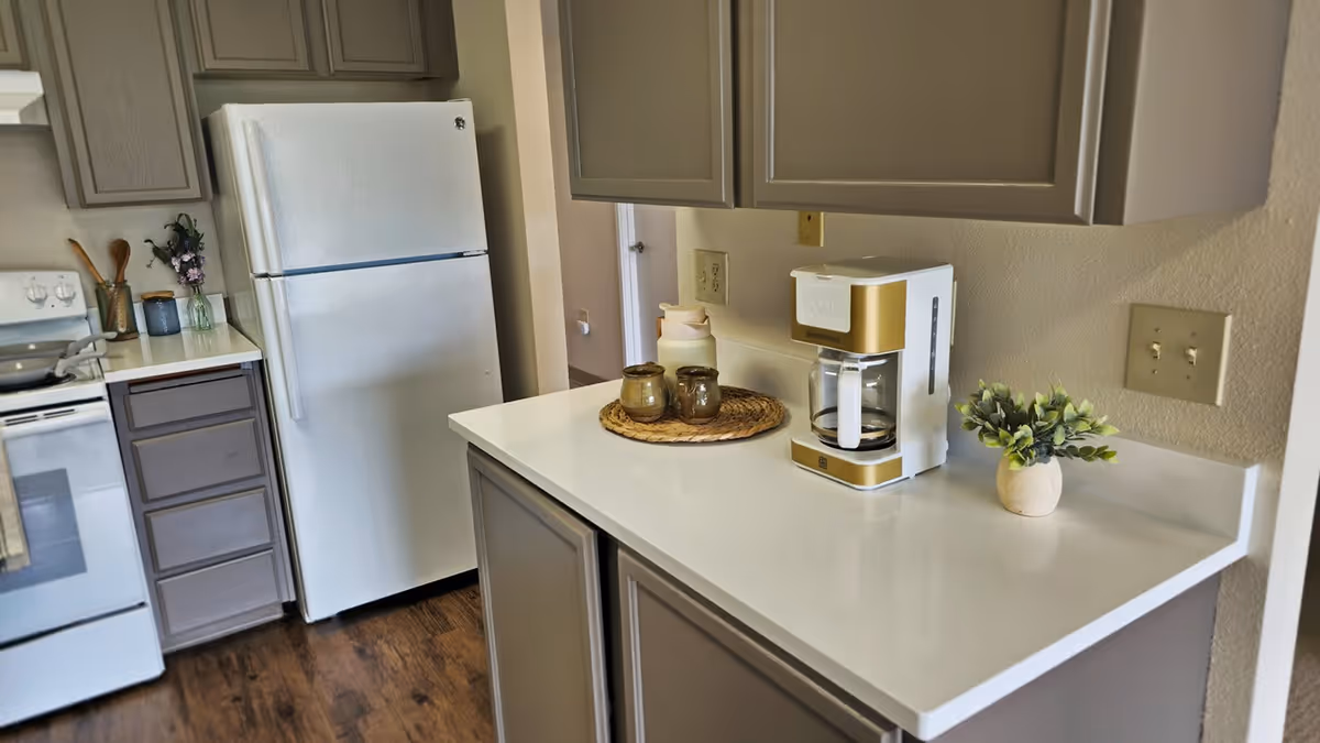 A kitchen area with gray cabinets, a white refrigerator, a white stove, and a white countertop. On the countertop, there is a coffee maker, a small plant in a pot, and a round woven placemat with two small jars. The floor is wooden.
