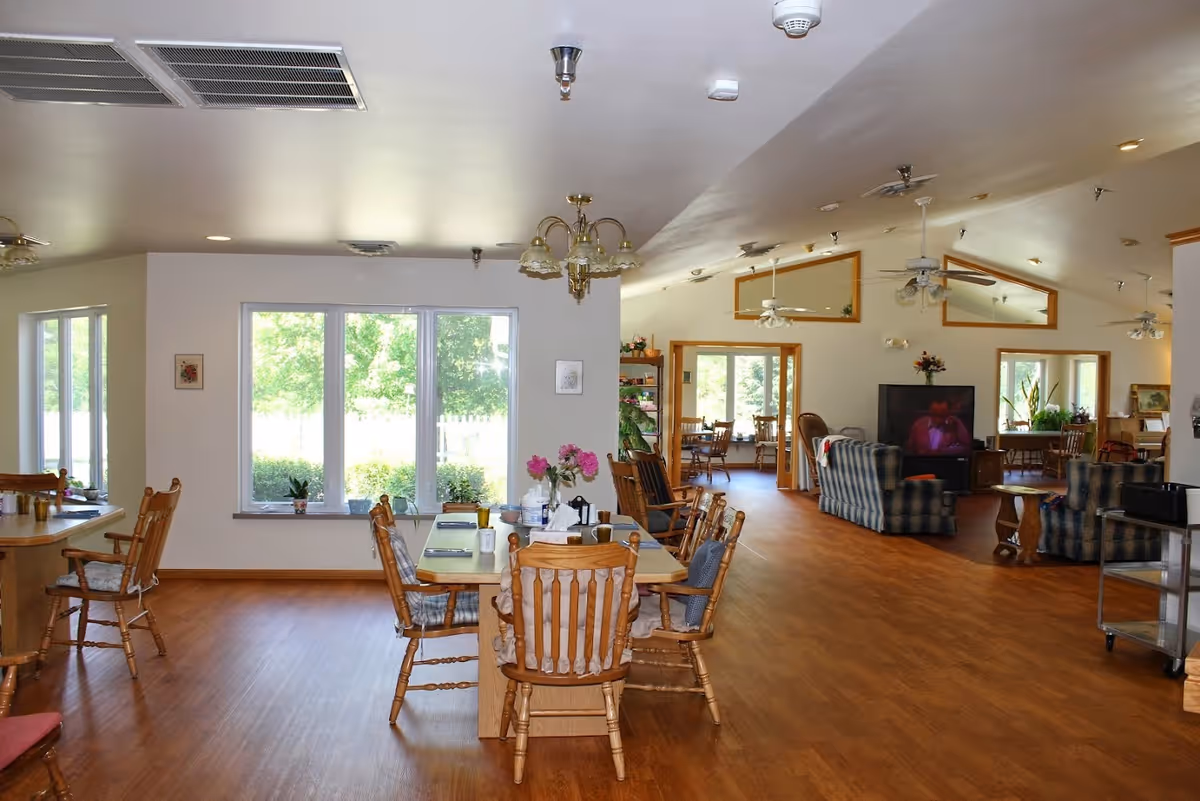 A spacious interior room in an assisted living facility featuring wooden floors, multiple wooden tables and chairs, large windows letting in natural light, and a seating area with plaid upholstered armchairs and a television. The room has ceiling fans and decorative wall mirrors, creating a comfortable and inviting atmosphere.