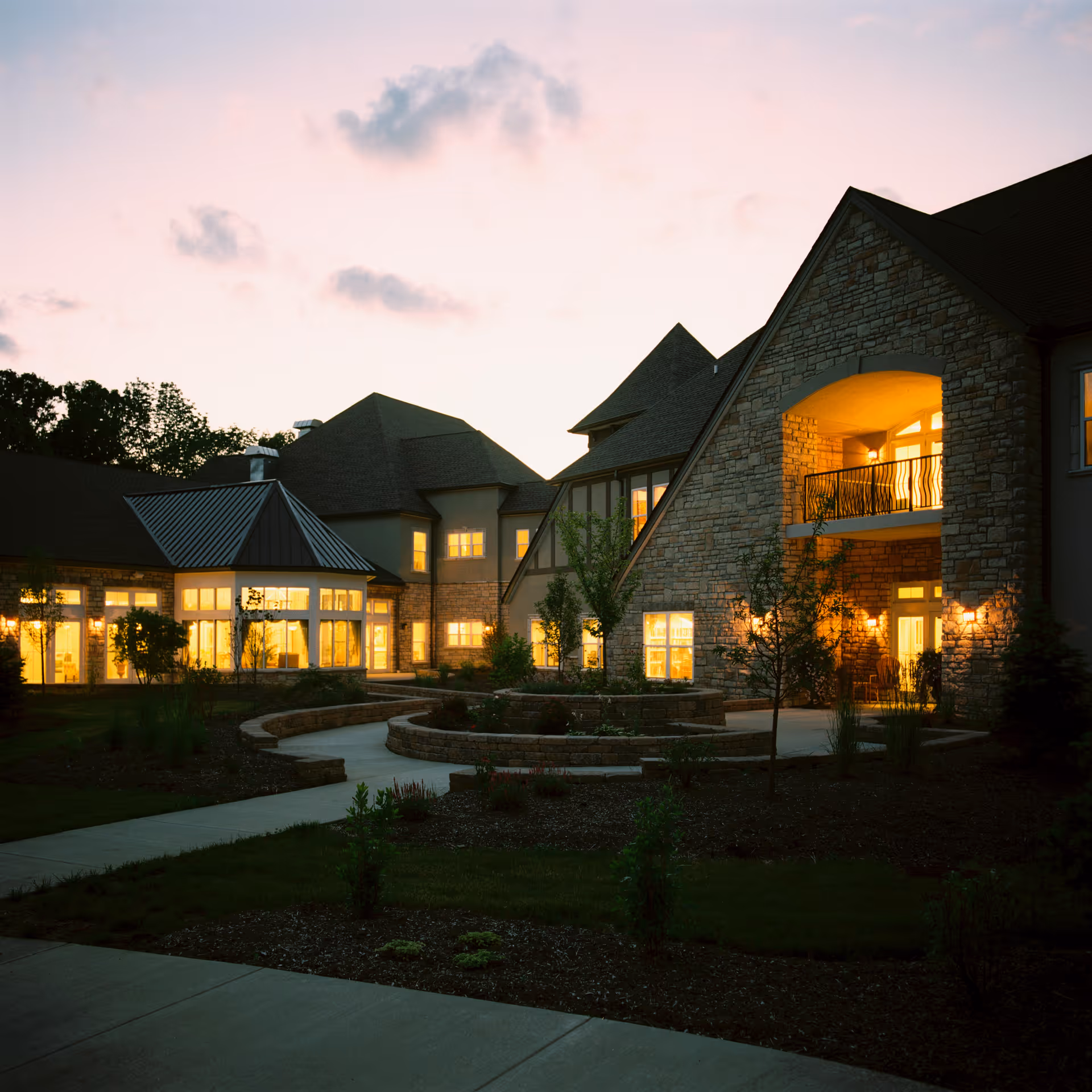 Exterior view of a stone-faced senior living building at dusk with illuminated windows and a curved landscaped walkway.