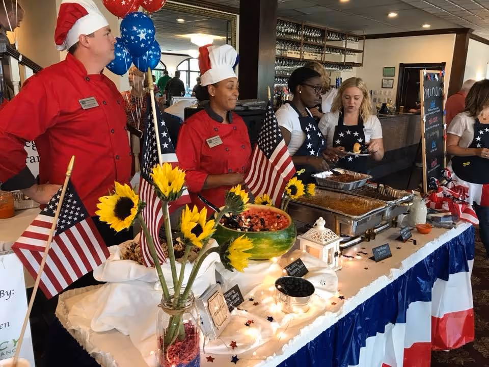 A festive buffet table decorated with American flags, sunflowers, and red, white, and blue decorations. Several staff members, including two chefs in red uniforms and white chef hats, and others wearing star-spangled aprons, are serving food behind the table in a dining area.