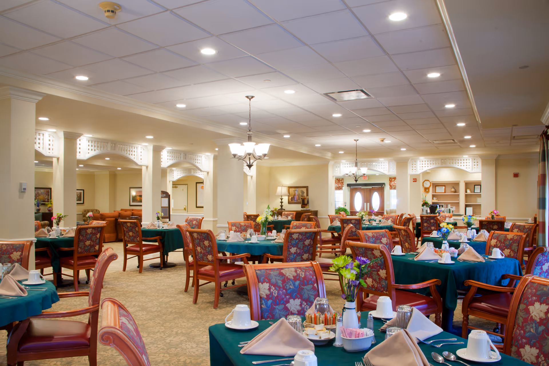 A spacious senior living dining room with tables set with green tablecloths, napkins, place settings, and floral centerpieces.