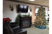 A cozy room decorated for Christmas featuring a lit Christmas tree with ornaments and a blue tree skirt, a fireplace with a garland on the mantle, a wall-mounted flat-screen TV above the fireplace, two dark leather chairs, and a poinsettia plant on the left side.