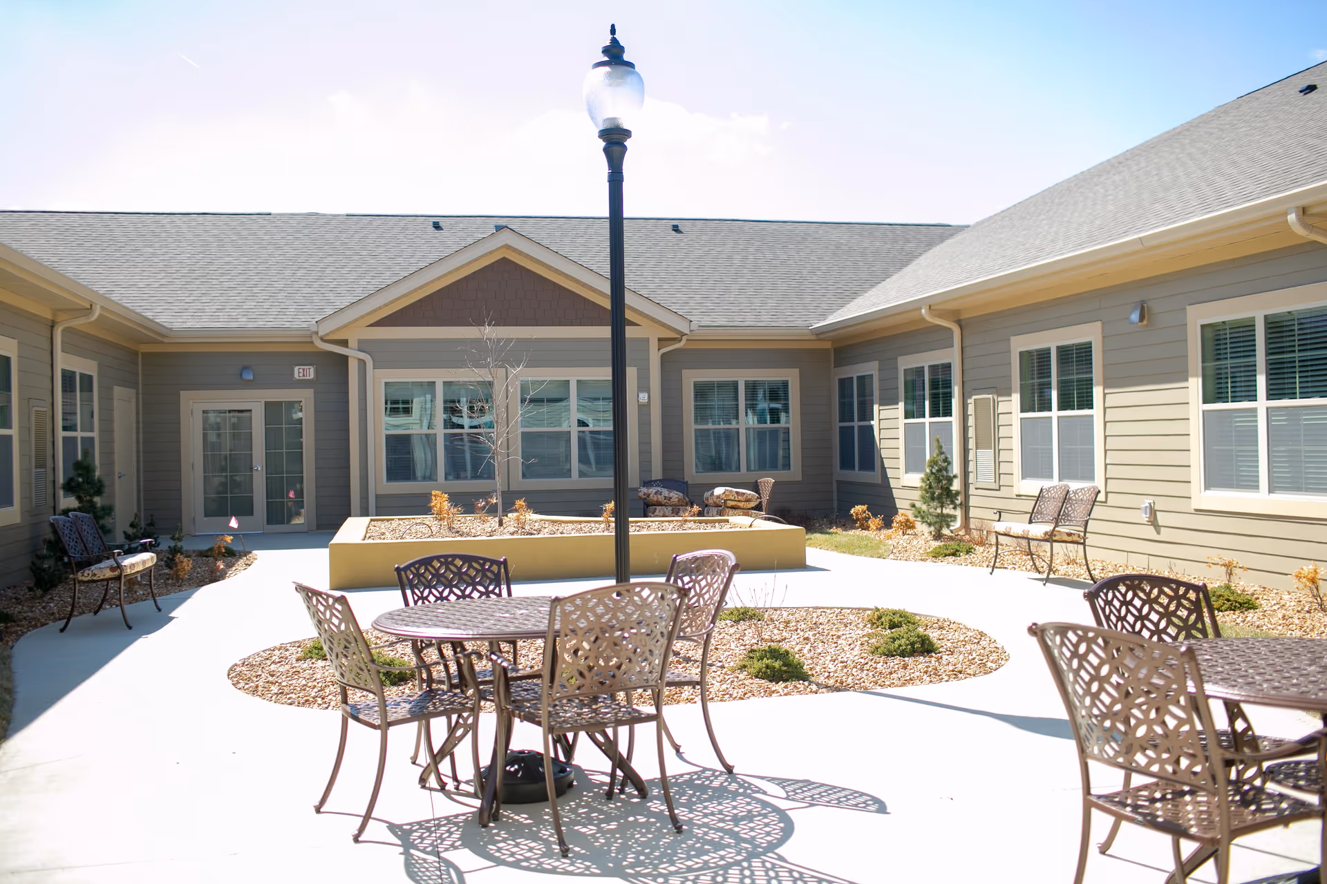 Outdoor courtyard area at The Boulevard Senior Living Of Wentzville with metal tables and chairs, a central lamp post, small landscaped garden beds with rocks and plants, surrounded by a single-story building with multiple windows and doors.