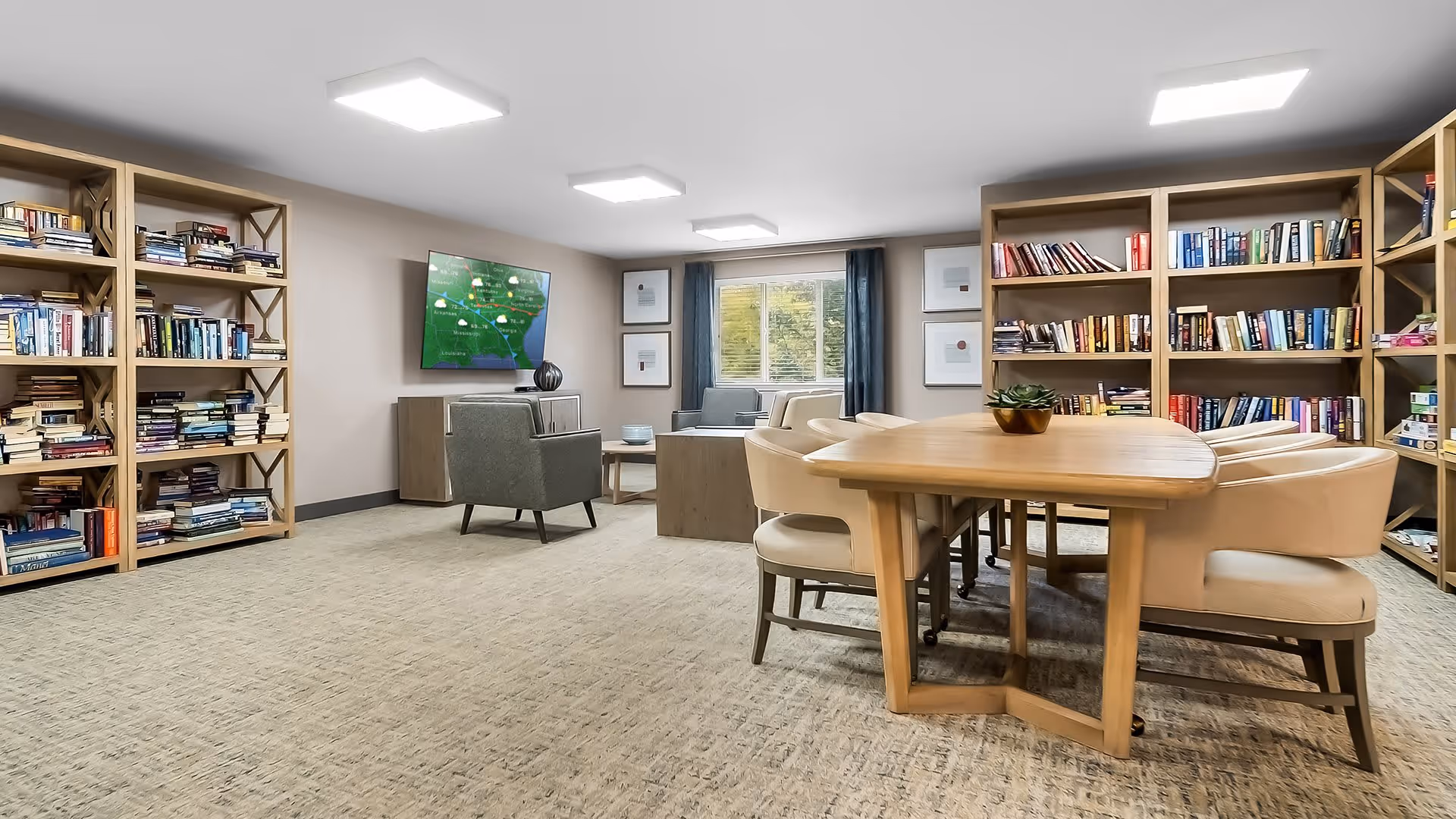 A cozy common room with bookshelves filled with books along the walls, a wooden table surrounded by cushioned chairs in the center, and a seating area with armchairs and a coffee table near a window. A flat-screen TV is mounted on the wall displaying a weather map.