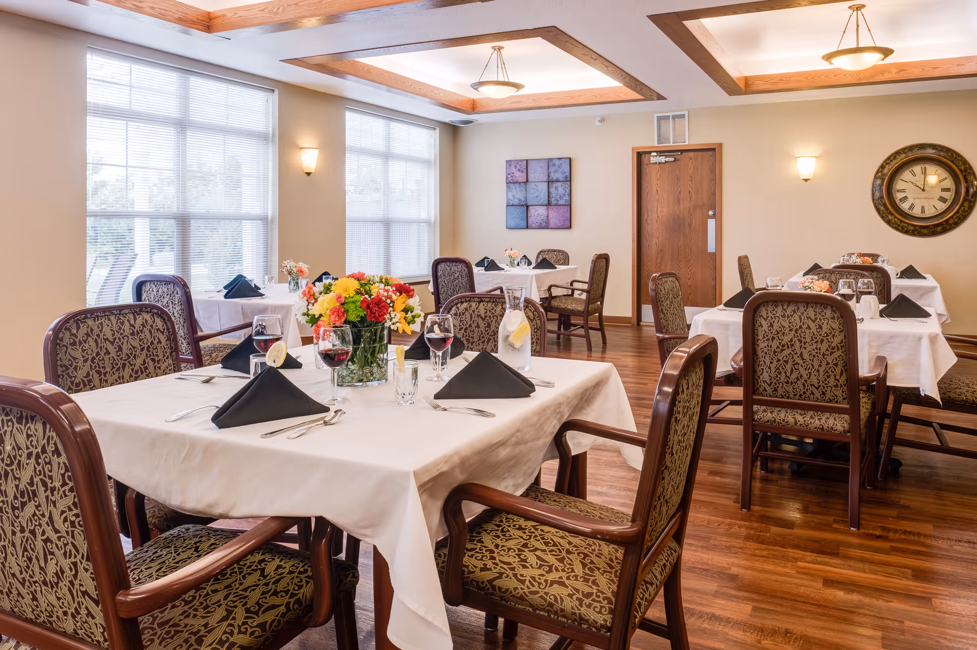 A well-lit dining room with several tables covered in white tablecloths, each set with black folded napkins, glasses of water and wine, and floral centerpieces. The room has large windows with blinds, wooden flooring, patterned upholstered chairs, and a wall clock. Ceiling lights and wall sconces provide additional lighting.