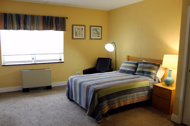 Furnished bedroom with a striped bed, wooden nightstand and lamp, black armchair, floor lamp, and a window.
