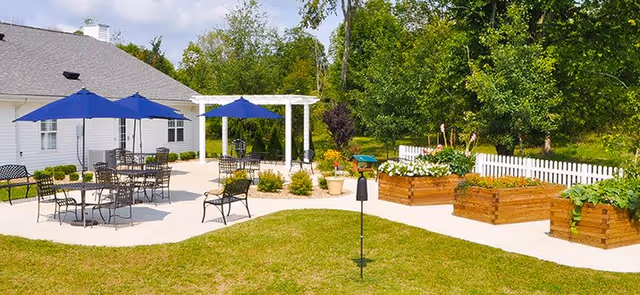 Outdoor patio area at The Meadows at Covington featuring multiple black metal tables and chairs with blue umbrellas, a white pergola, raised wooden garden beds with flowers and plants, a white picket fence, and a grassy lawn surrounded by trees.
