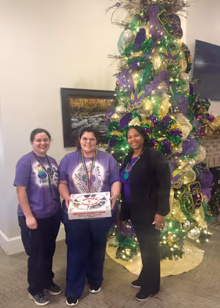 Three women standing indoors in front of a decorated Christmas tree with purple, green, and gold ornaments and ribbons. The woman in the middle is holding a box of Krispy Kreme doughnuts. They are smiling and wearing casual clothing, with two of them in purple shirts. A framed picture and a television mounted on the wall are visible in the background.