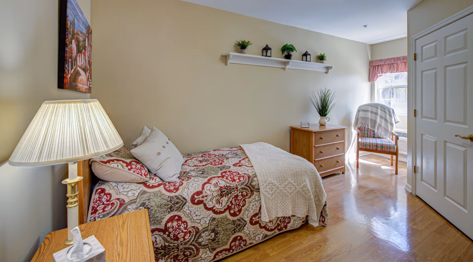 A cozy bedroom with a single bed covered in a patterned red, beige, and green bedspread with a white knitted throw blanket. There is a wooden nightstand with a lamp and a tissue box next to the bed. On the wall above the bed is a painting, and a white shelf with small plants and lanterns is mounted on the opposite wall. A wooden dresser with a plant on top is near a window with pink valance curtains. A wooden chair with a striped cushion and a white throw blanket is positioned by the window. The floor is polished wood, and a closed white door is visible on the right.