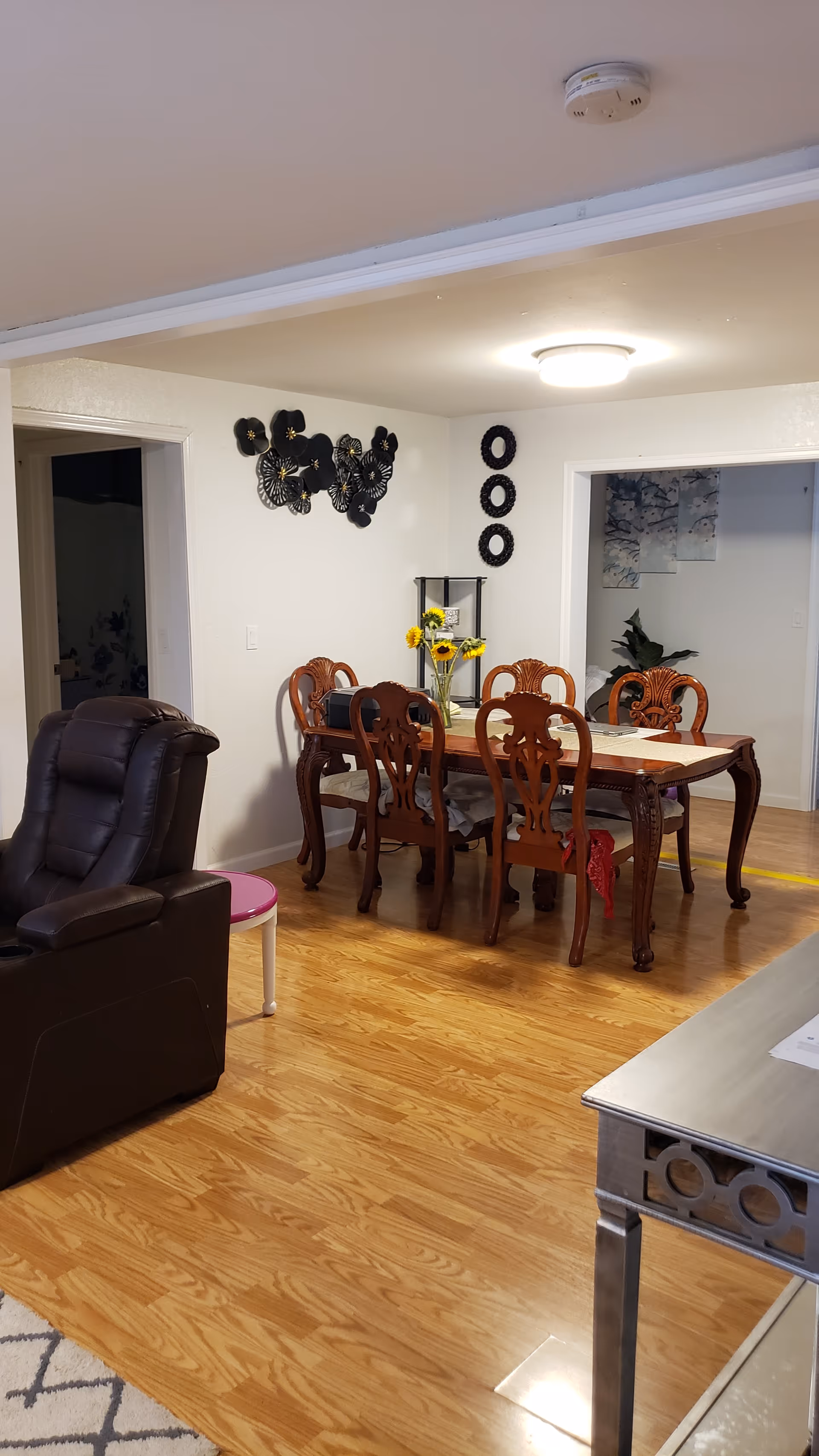 Interior view of a dining area in a senior living facility with a wooden dining table and six matching chairs. The table has a vase with sunflowers and a table runner. There is a dark brown recliner chair and a small pink and white side table nearby. The walls are decorated with black circular and floral wall art. The floor is wooden, and there is a ceiling light fixture.