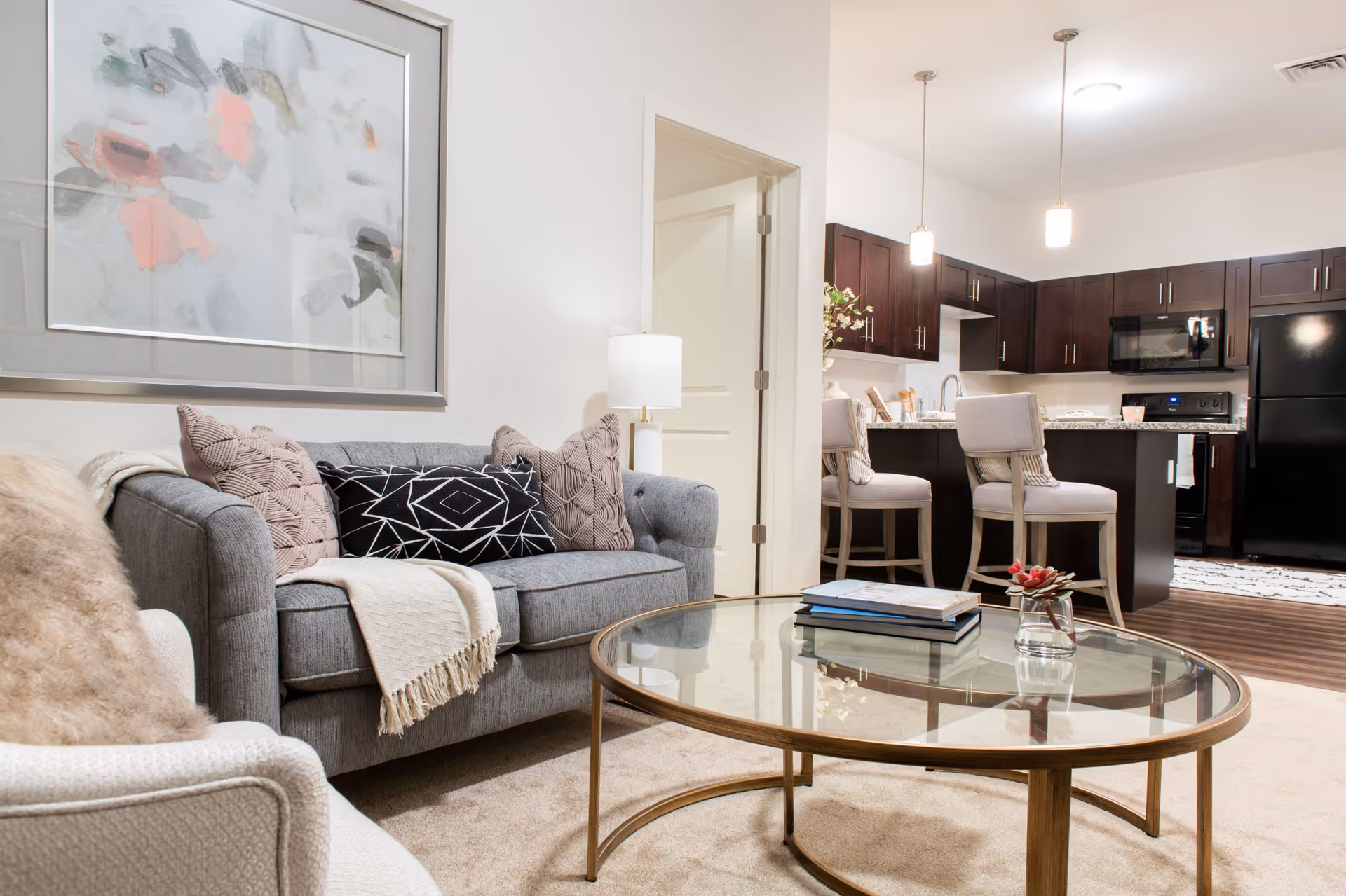 A modern living room with a gray sofa adorned with decorative pillows and a throw blanket, a glass coffee table with books and a small plant, and a view into a kitchen area with dark wood cabinets, a black refrigerator, stove, and two bar stools at a kitchen island.