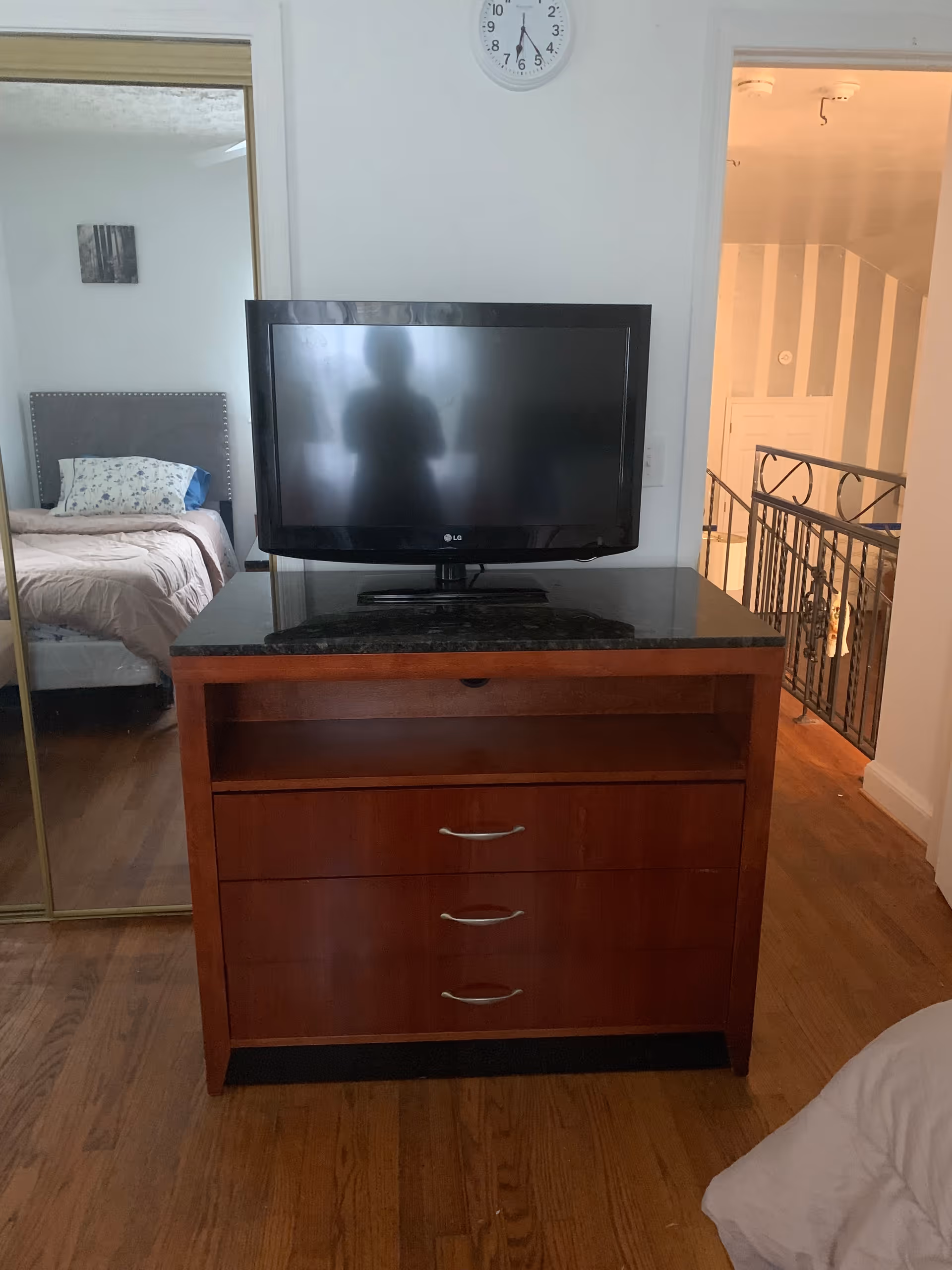 A bedroom interior featuring a wooden dresser with a black granite top, on which a flat-screen LG television is placed. Behind the dresser, a wall clock shows the time as 6:00. To the left, a mirrored sliding closet door reflects a bed with a gray upholstered headboard and floral pillows. To the right, an open doorway reveals a hallway with a decorative railing and striped wallpaper.