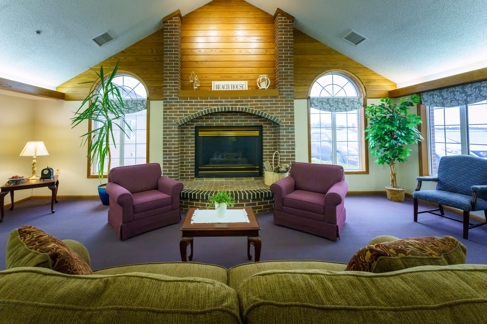 A cozy living room with a brick fireplace centered on a wooden paneled wall. Two purple armchairs face the fireplace with a small wooden coffee table between them, topped with a white doily and a small potted plant. A green sofa with patterned cushions is in the foreground. Large arched windows with floral valances flank the fireplace, letting in natural light. There are two tall plants in pots near the windows, and a side table with a lamp and clock on the left side of the room.