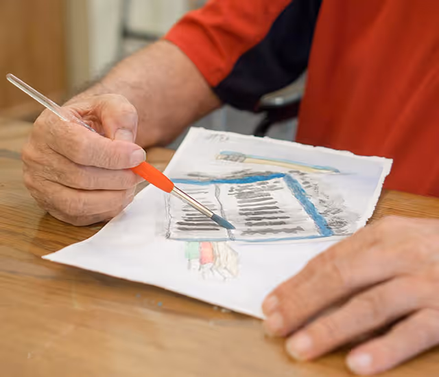 Close-up of a person painting a watercolor picture on a piece of paper placed on a wooden table. The person is holding a paintbrush with a red handle and is wearing a red shirt.