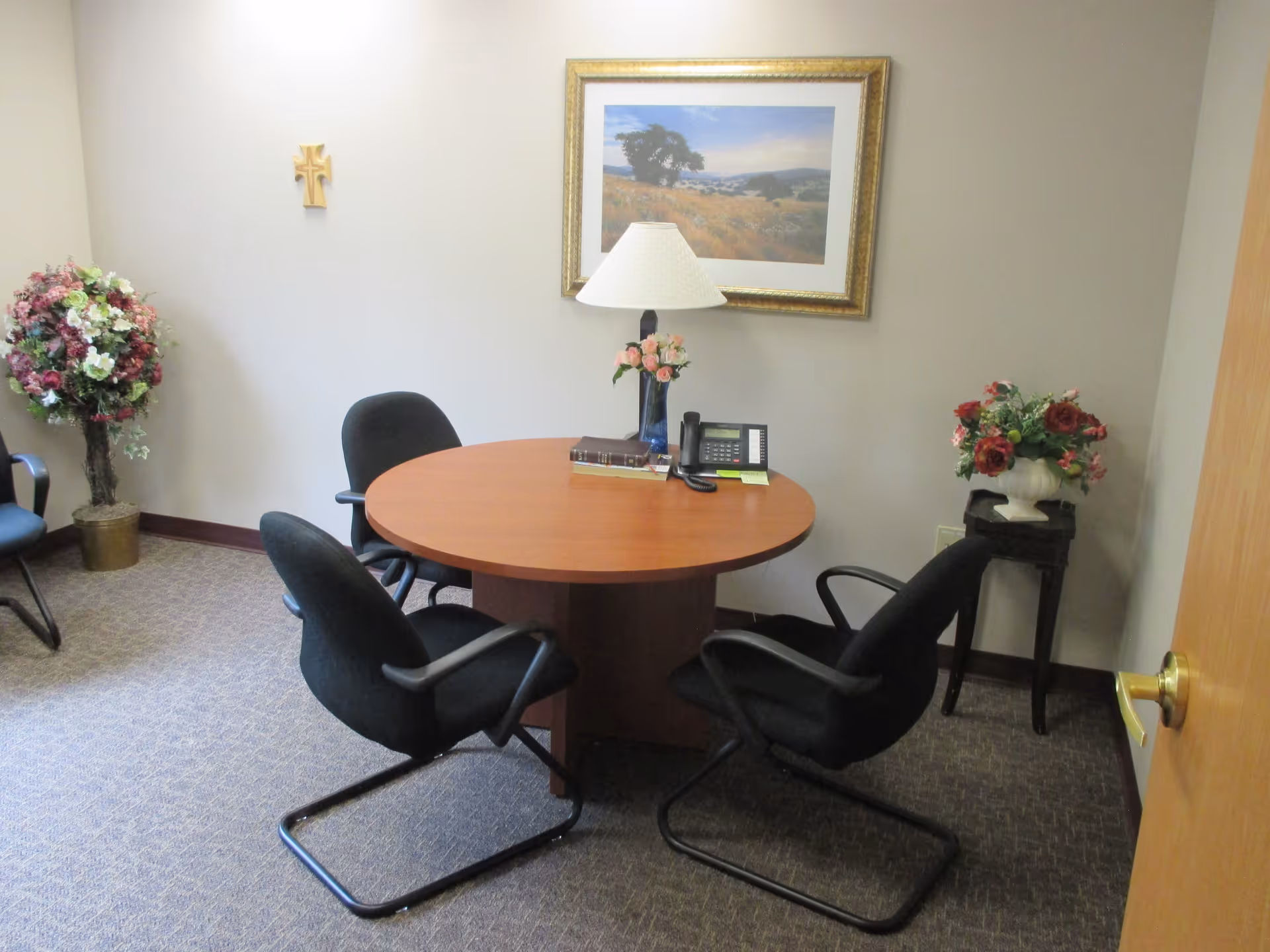 A small office or meeting room with a round wooden table surrounded by four black office chairs. On the table are a lamp, a telephone, a book, and a small vase with flowers. The room has beige walls, a framed landscape painting, a wooden cross on the wall, and two flower arrangements in pots placed on the floor and a small stand.