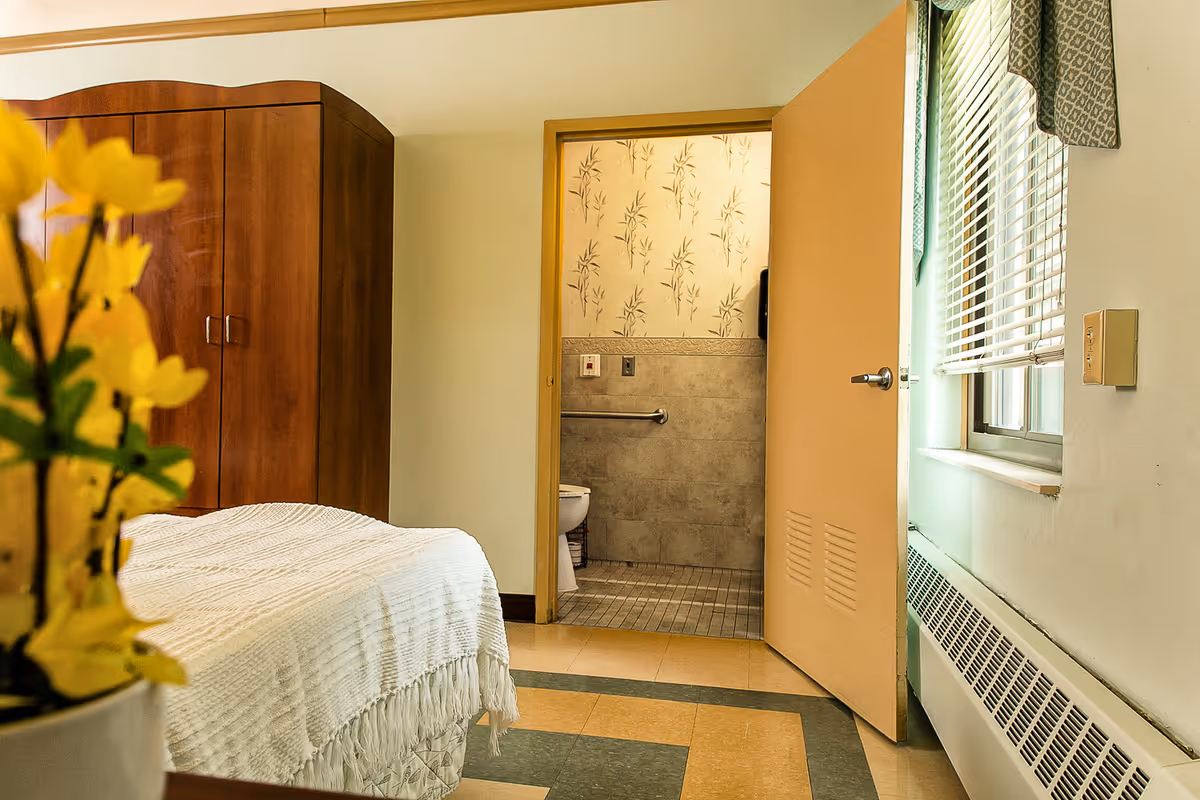 A cozy bedroom in a senior living facility with a bed covered in a white textured blanket, a wooden wardrobe, and a vase with yellow flowers in the foreground. The door to an adjacent bathroom is open, revealing a toilet and tiled walls with a floral pattern. A window with blinds and a patterned valance lets in natural light.