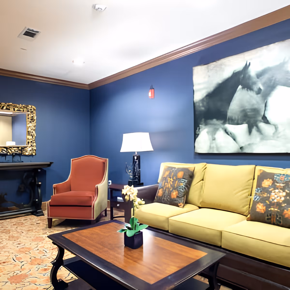A cozy living room with a yellow sofa adorned with decorative pillows, a red armchair, a wooden coffee table with a small potted plant, a table lamp, and a large black and white painting of horses on a blue wall.