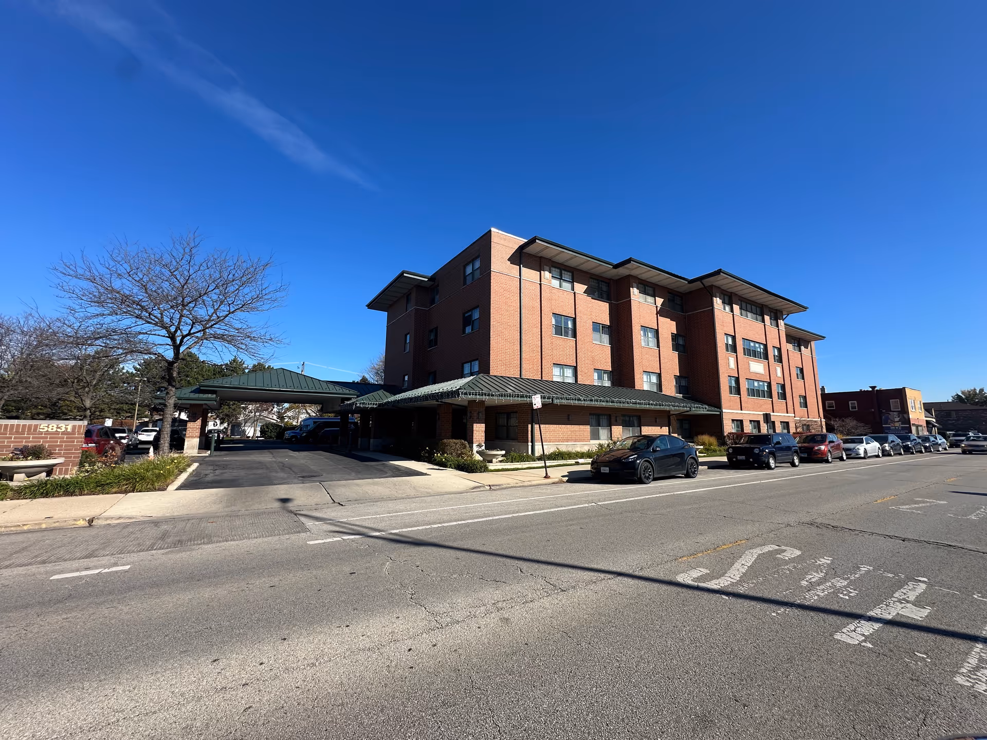 Exterior view of a four-story brick building with a green roof canopy over the entrance, several parked cars along the street, a leafless tree, and a clear blue sky.