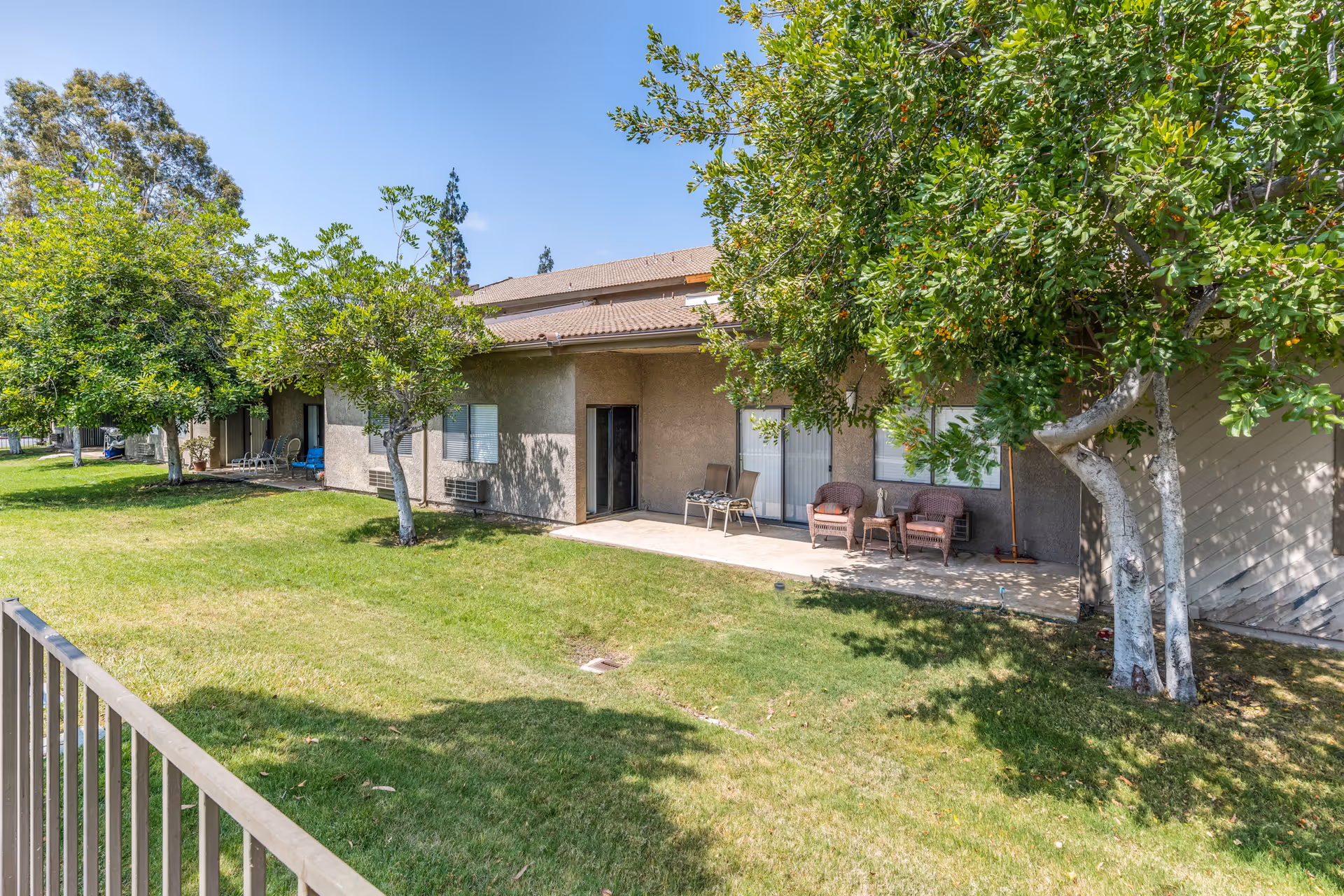 Outdoor view of a single-story building with a patio area featuring chairs and a small table. The building is surrounded by green grass and several trees under a clear blue sky.