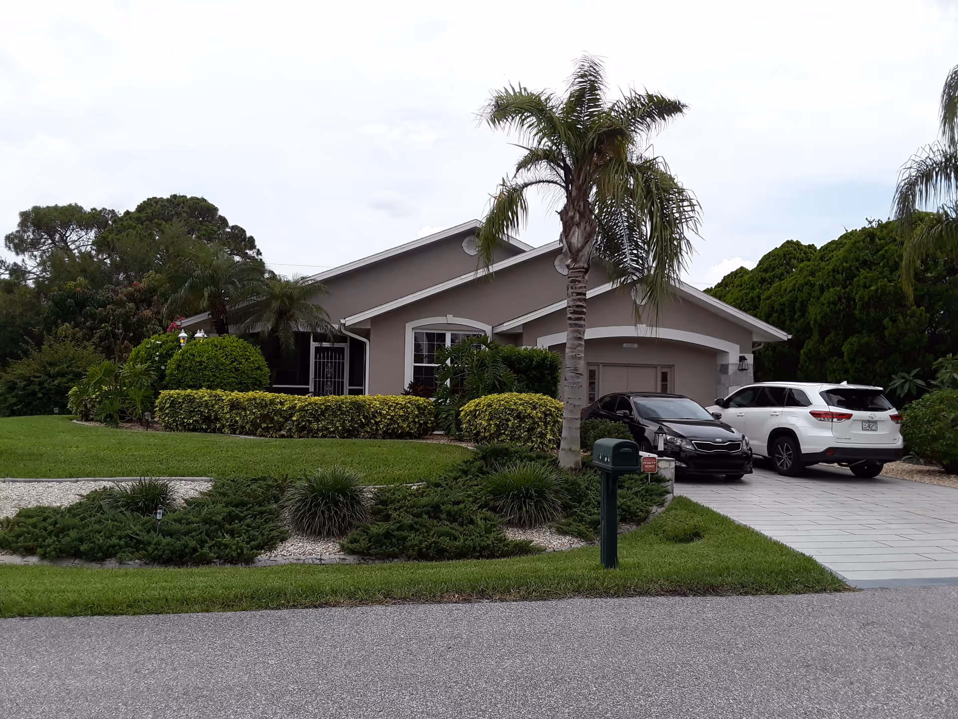 A single-story house with a well-maintained front yard featuring green grass, bushes, and palm trees. Two cars are parked in the driveway, and a green mailbox is visible near the street.