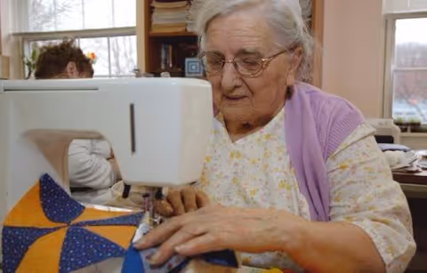An elderly woman with glasses and a lavender shawl is using a sewing machine to stitch a colorful quilt in a well-lit room with windows and shelves in the background.
