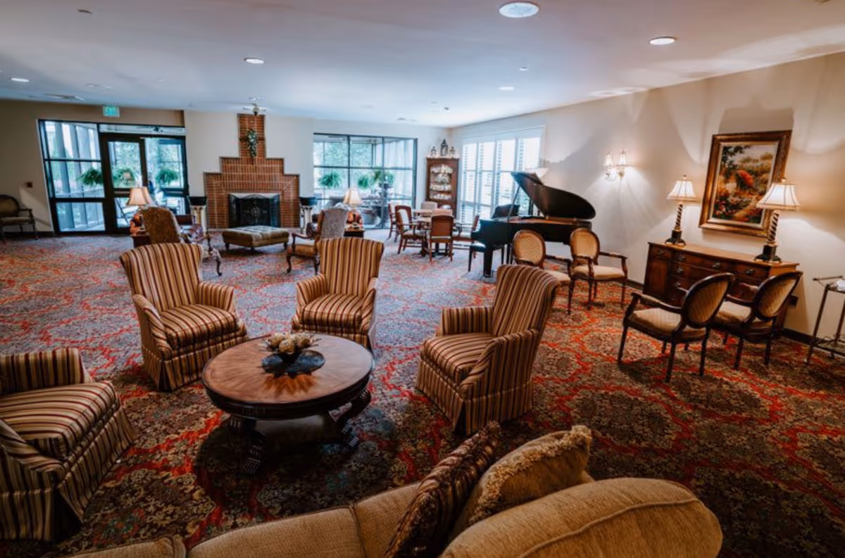 A spacious and elegantly furnished living room with patterned carpet, several striped armchairs arranged around a round wooden coffee table, a grand piano near the wall, a fireplace with a brick surround, and large windows letting in natural light. The room also features wall lamps, framed artwork, and additional seating areas.