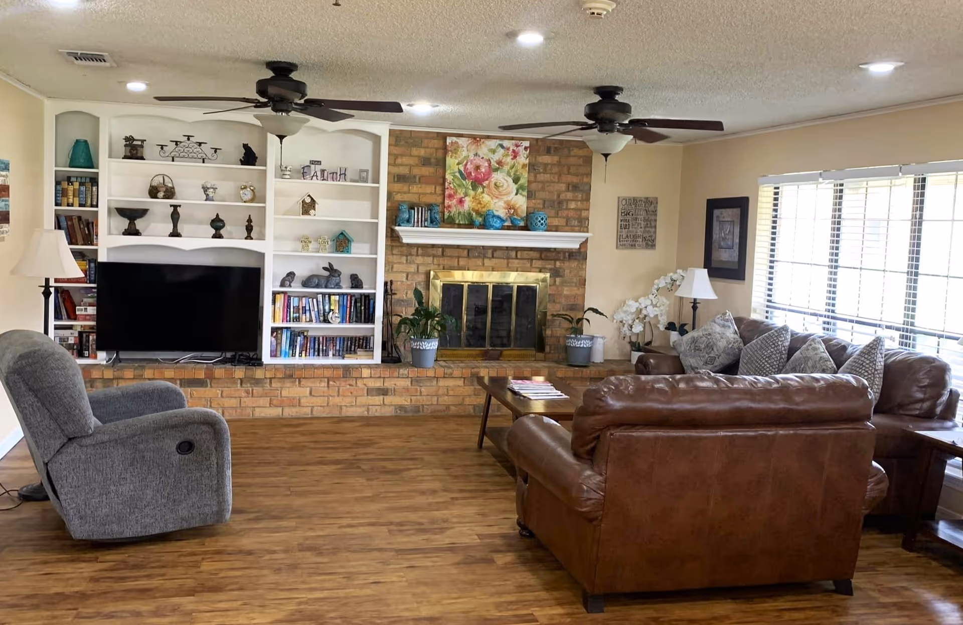 A cozy living room with wooden flooring, a brick fireplace with a floral painting above it, built-in white shelves filled with books and decorative items, a flat-screen TV, a gray recliner chair, and brown leather sofas with patterned cushions. Two ceiling fans with lights are mounted on the ceiling, and large windows with blinds allow natural light to fill the room.
