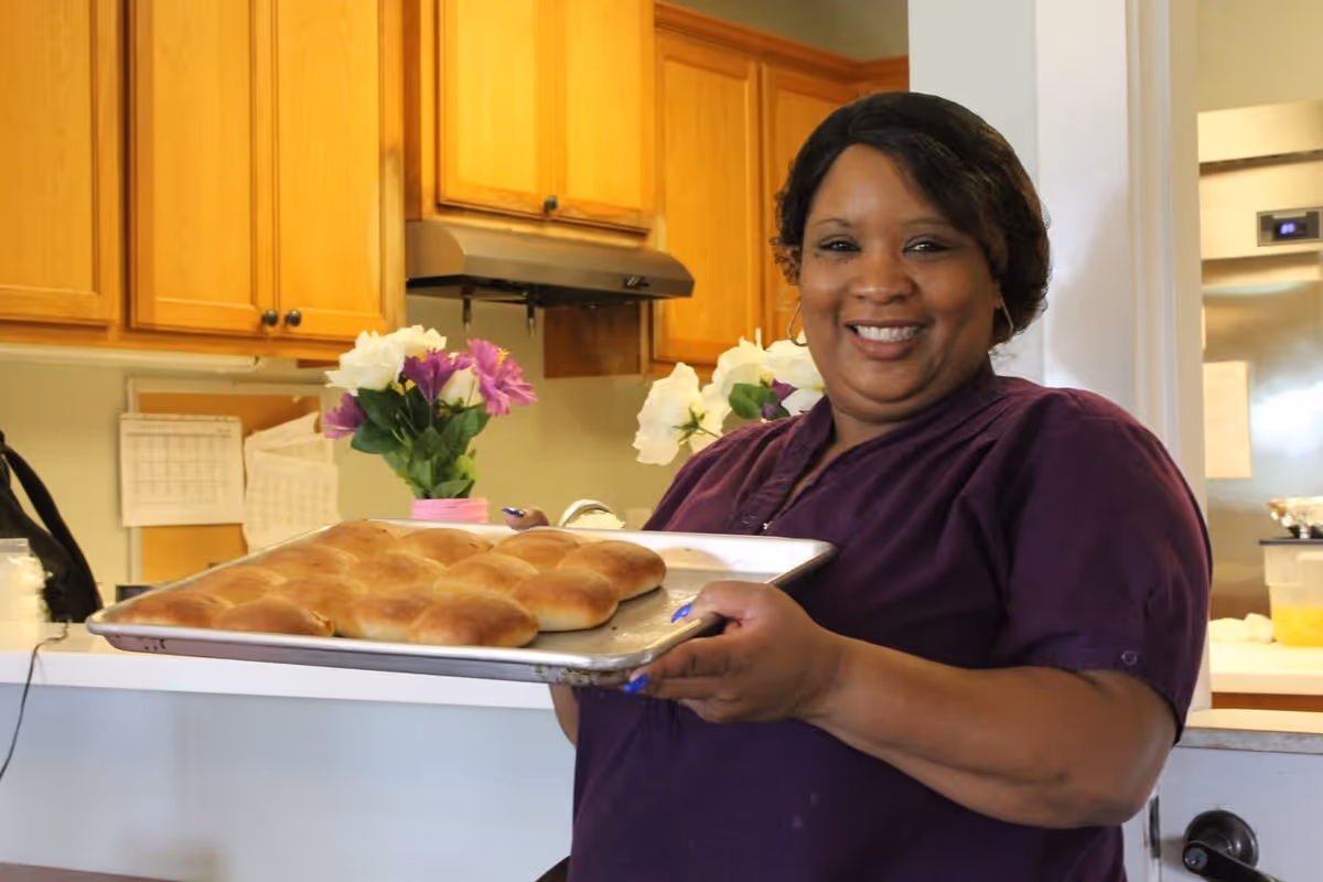 A smiling woman in a purple shirt holding a baking tray with freshly baked bread rolls in a kitchen setting with wooden cabinets and flowers in the background.