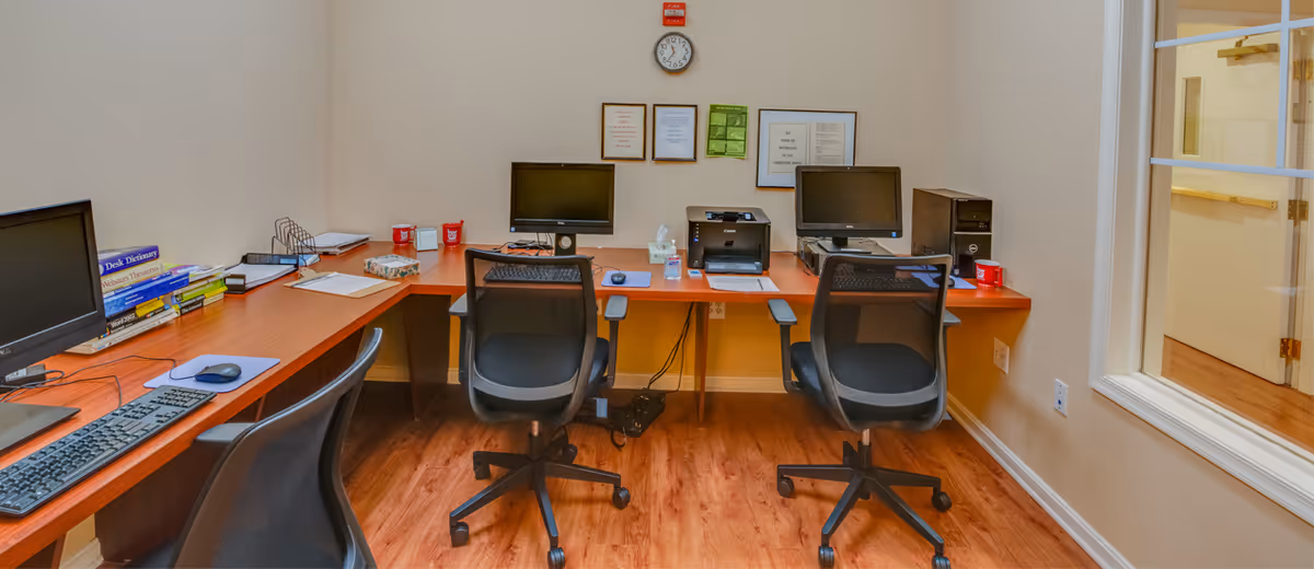 A small office area with a wooden L-shaped desk against the walls, three black mesh office chairs, two computer monitors, a keyboard, a printer, office supplies, and framed certificates on the wall above the desk. The room has light-colored walls and wood flooring, with a window looking into an adjacent hallway.