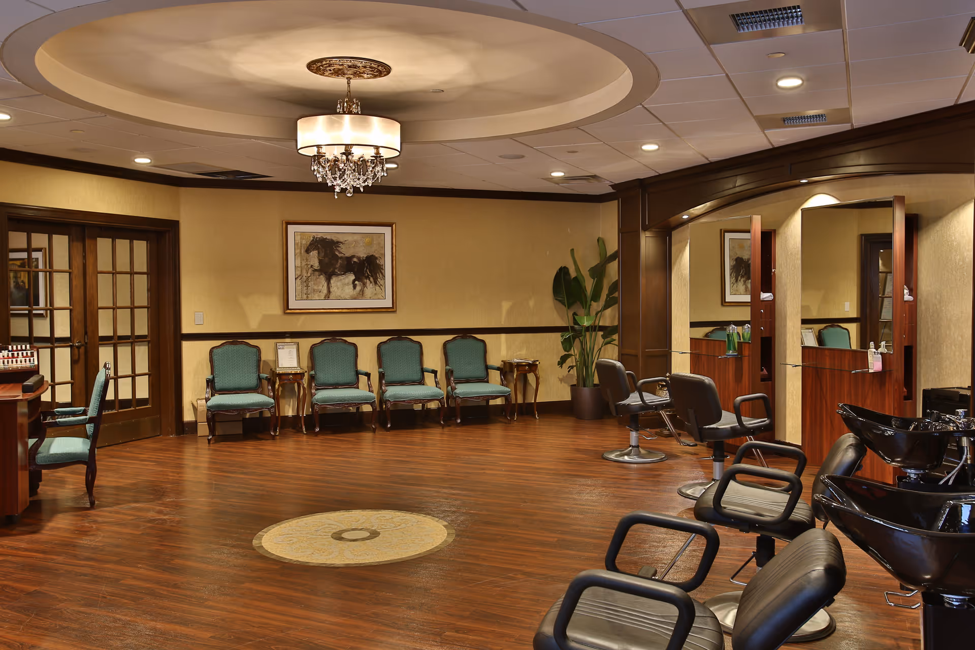 Interior of a salon area in a senior living facility with wooden flooring, a circular decorative ceiling feature with a chandelier, green upholstered chairs lined up against the wall, a framed horse painting, and salon stations with black styling chairs and wash basins.
