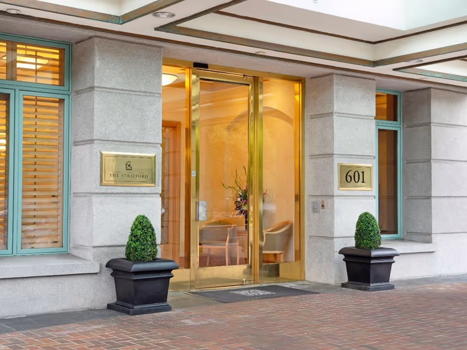 Entrance to The Stratford facility with glass double doors framed in gold, two potted plants on either side, and plaques displaying the name 'The Stratford' and the number '601'. Windows with green frames and shutters flank the entrance.