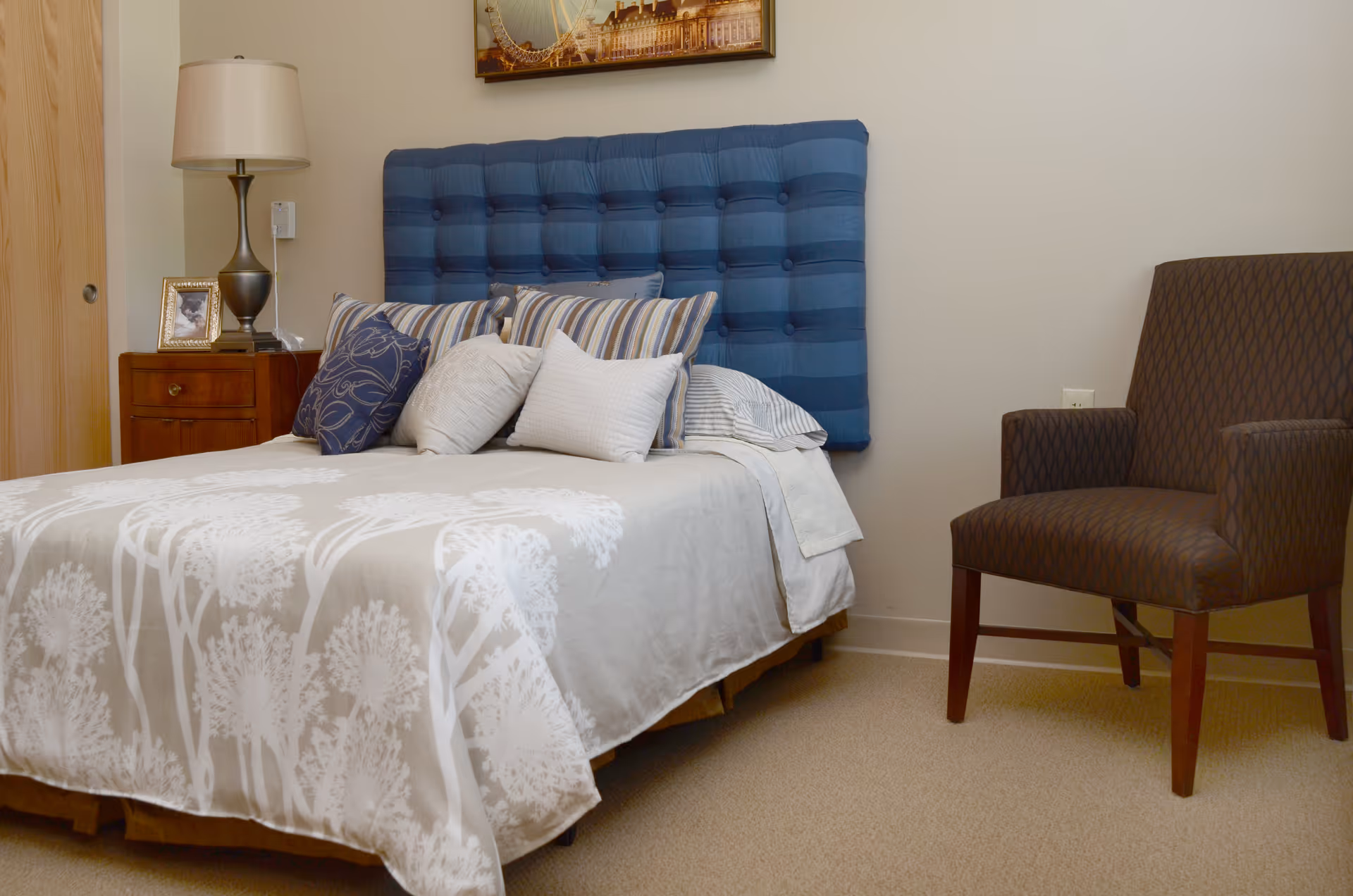 Neatly made bed with a blue tufted headboard, striped and patterned pillows, a nightstand with a lamp and framed photo, and an upholstered chair in a small bedroom.