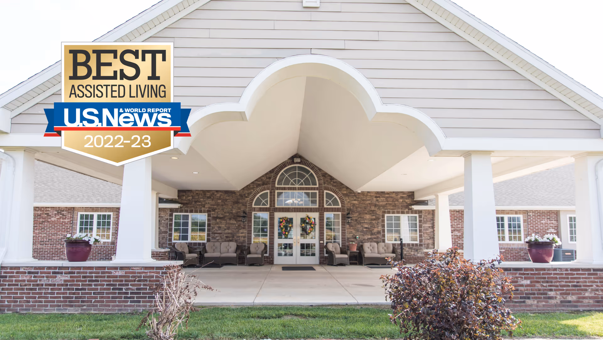 Covered front entrance of a senior living building with seating, planters, and a decorative archway, with an award badge in the top-left.