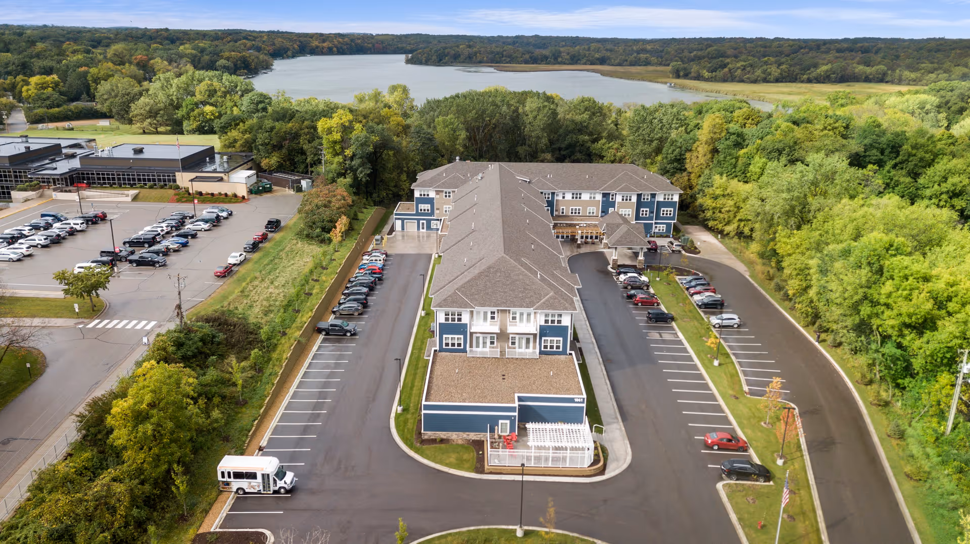 Aerial view of Harrison Bay Senior Living facility showing a large, multi-story building with a gray roof and blue and beige exterior. The building is surrounded by parking lots with numerous cars parked, and a small shuttle bus is visible in one parking spot. The facility is bordered by lush green trees and overlooks a large body of water with a forested area in the background under a partly cloudy sky.