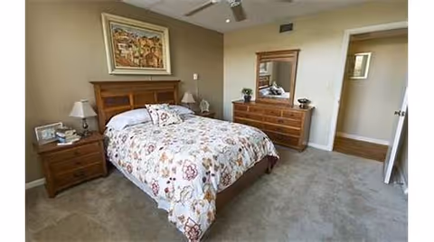 A bedroom with a wooden bed frame and a floral patterned bedspread. There are two wooden nightstands on either side of the bed, each with a lamp. A wooden dresser with a mirror is against the wall, and a framed picture hangs above the bed. The room has beige walls and carpeted flooring.